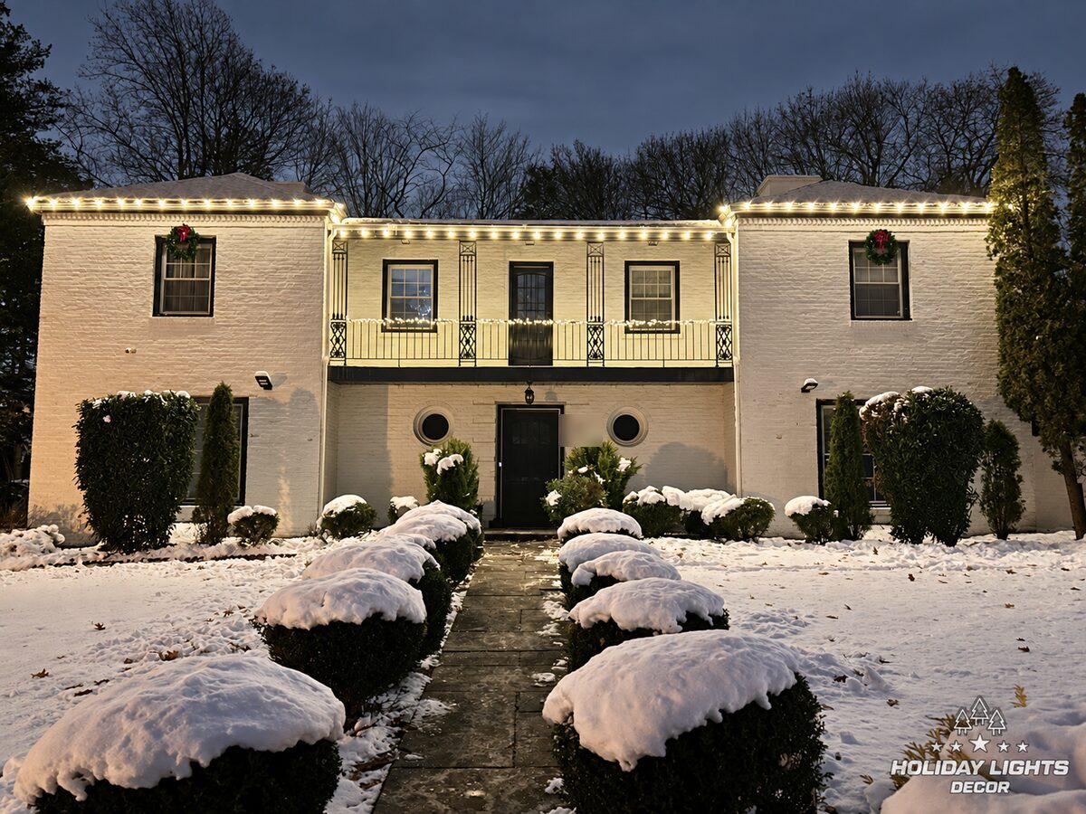 White-painted brick home in Orange with warm-white roofline and balcony lights, two window wreaths and snow.