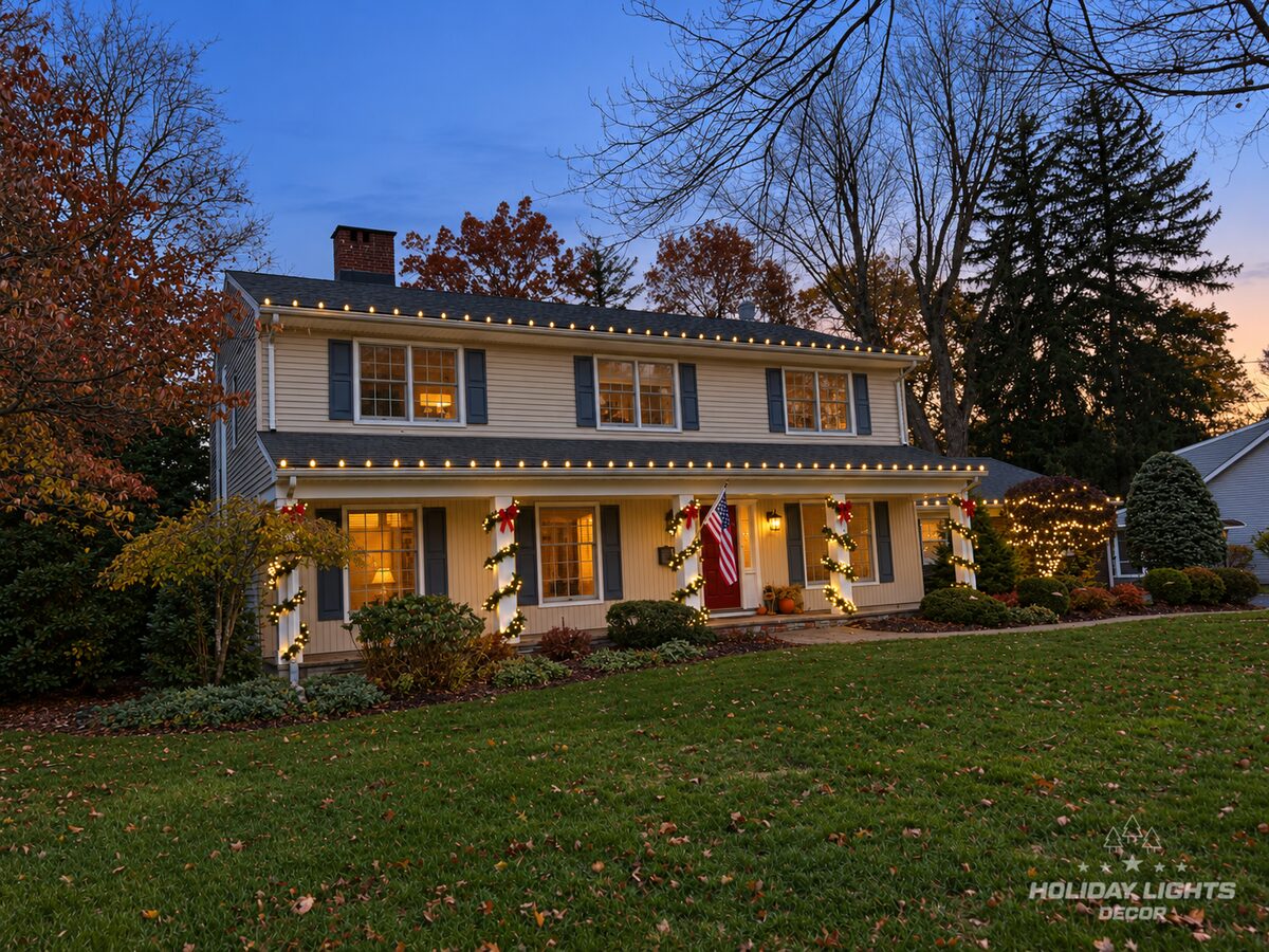 Angled view of a colonial at dusk with warm-white roofline lights and lit garland on the porch columns.
