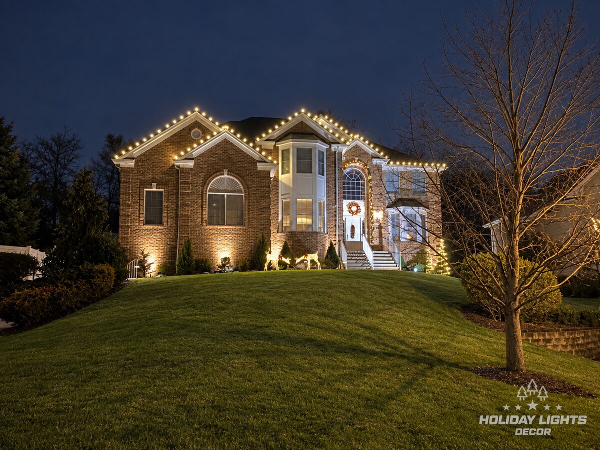 Brick home in Alpine at night with warm-white lights on two front gables, a lit entry arch, wreath and lit reindeer.